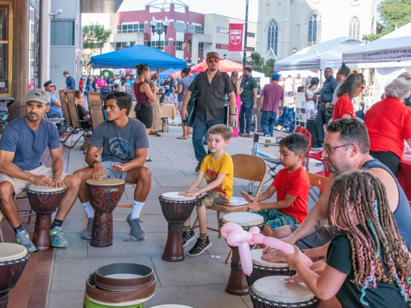 Children and adults sitting in a circle on the sidewalk playing drums